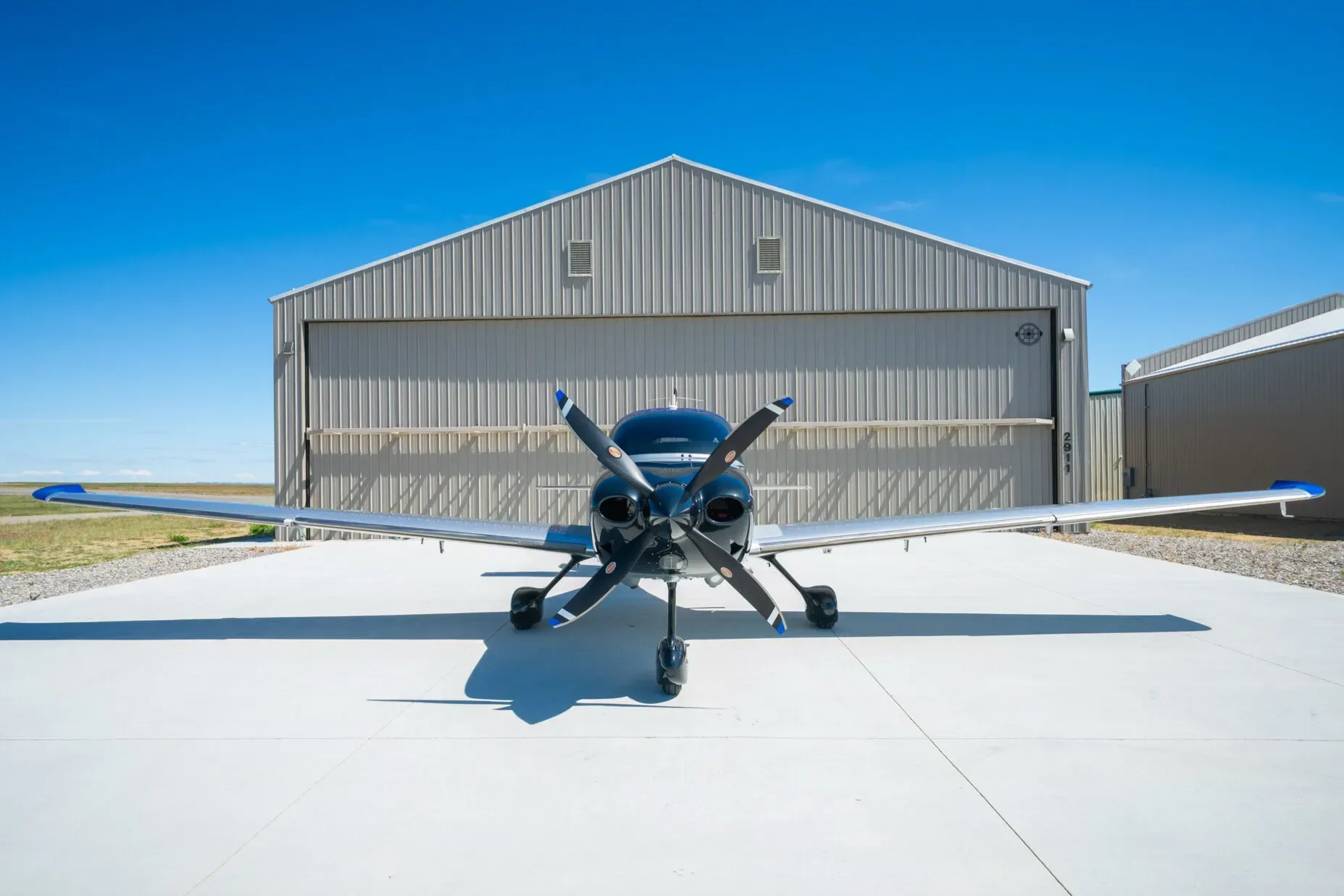 A front view of the Cirrus SR22T aircraft with a silver fuselage and black propeller, parked in front of a hangar.