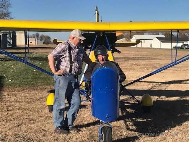 A yellow and blue ultralight aircraft on a grassy field with two men standing next to it.