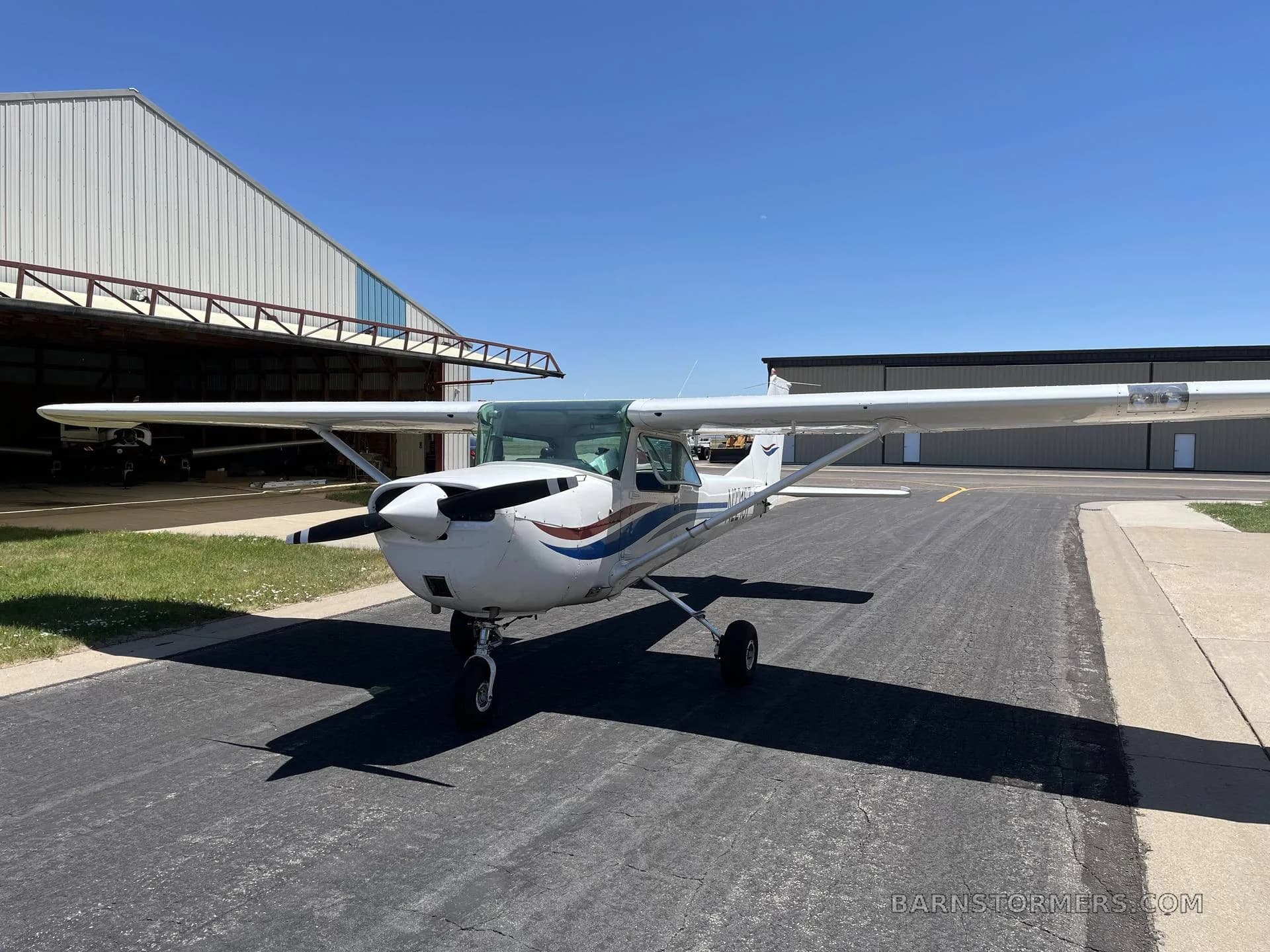 A white Cessna 150/152 type aircraft with blue and red stripes parked on an asphalt ramp in front of a hangar on a sunny day.