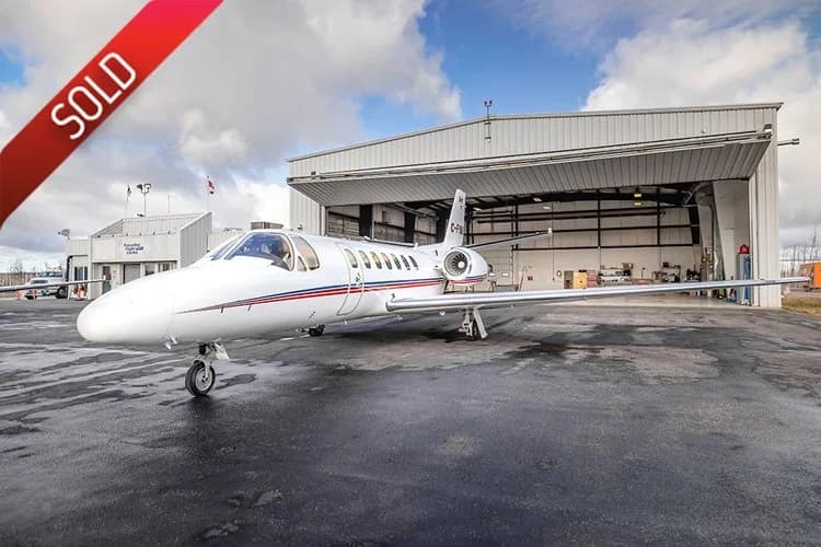 A white Cessna Citation jet with red and blue stripes parked on a wet tarmac in front of a hangar.