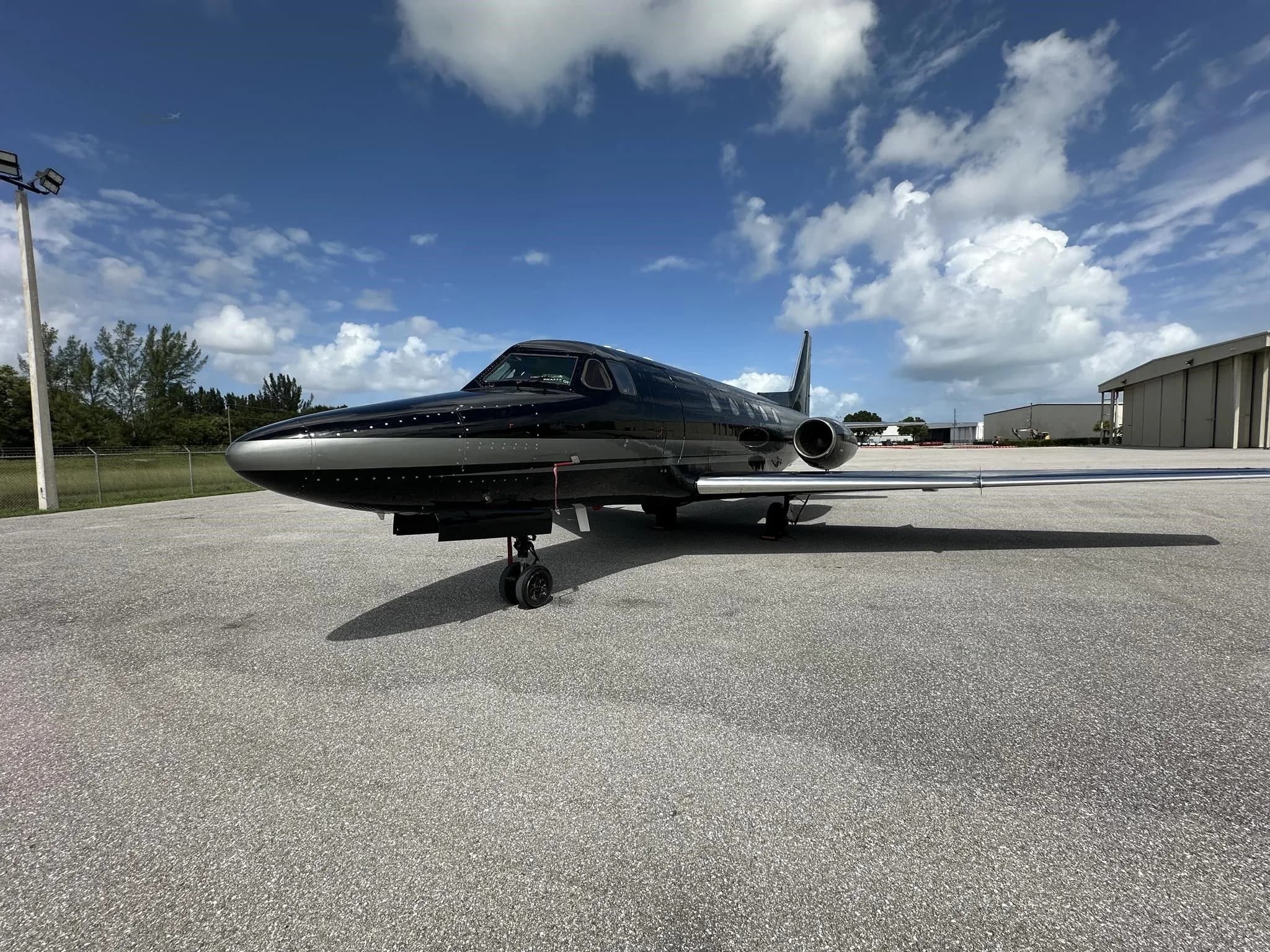 A front-side view of a black and silver business jet parked on an asphalt tarmac under a bright blue sky.