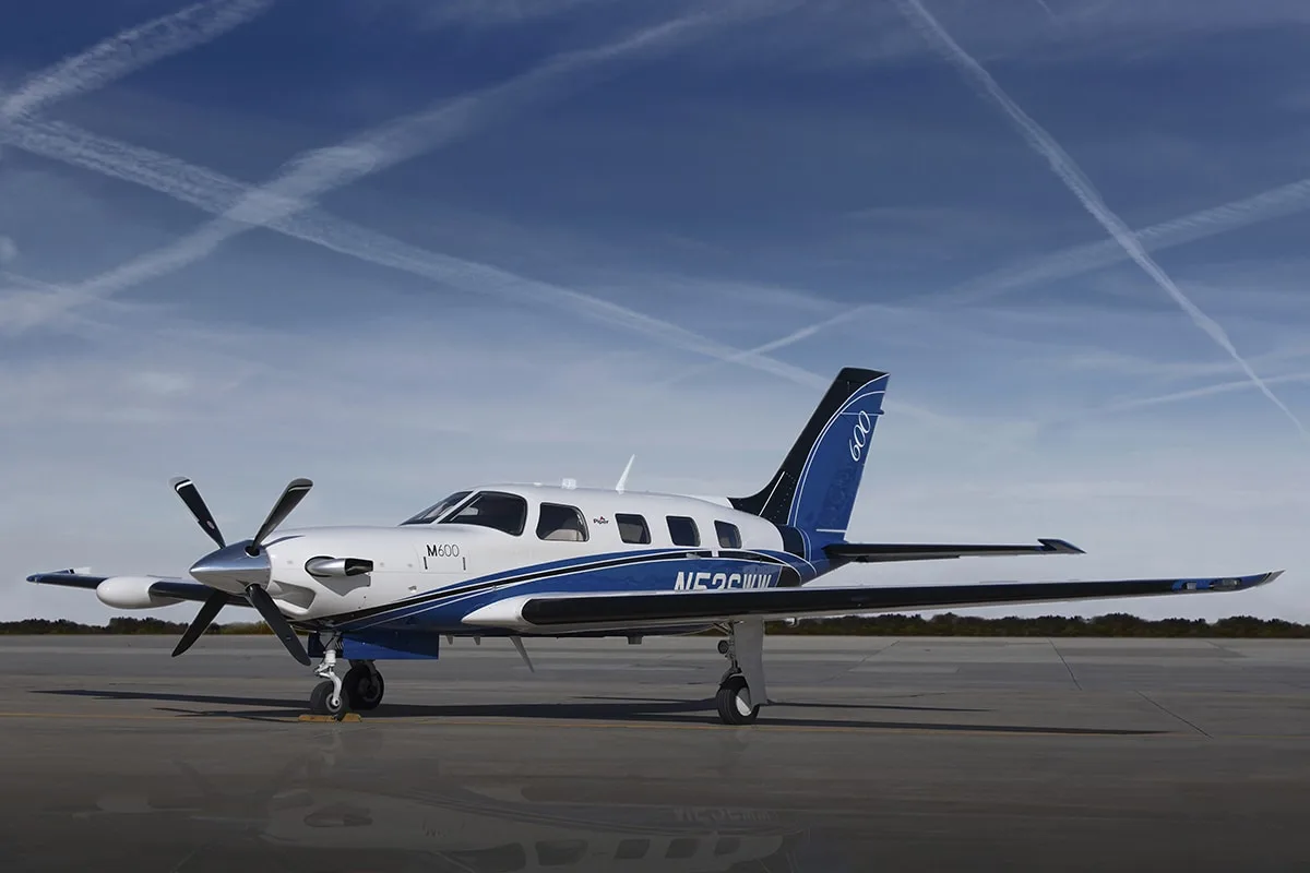 A white Piper M600 turboprop aircraft with blue and black stripes is parked on a tarmac under a blue sky with contrails.