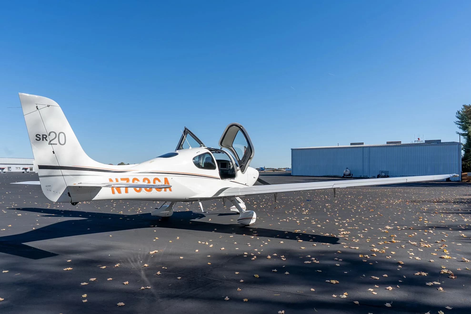 A white Cirrus SR20 aircraft with its doors open is parked on an asphalt tarmac, viewed from the rear right.