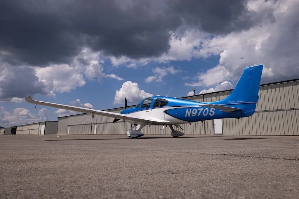 A low-angle rear-side view of the blue and white Cirrus SR22 aircraft, N970S, parked on an asphalt ramp with hangars in the background.