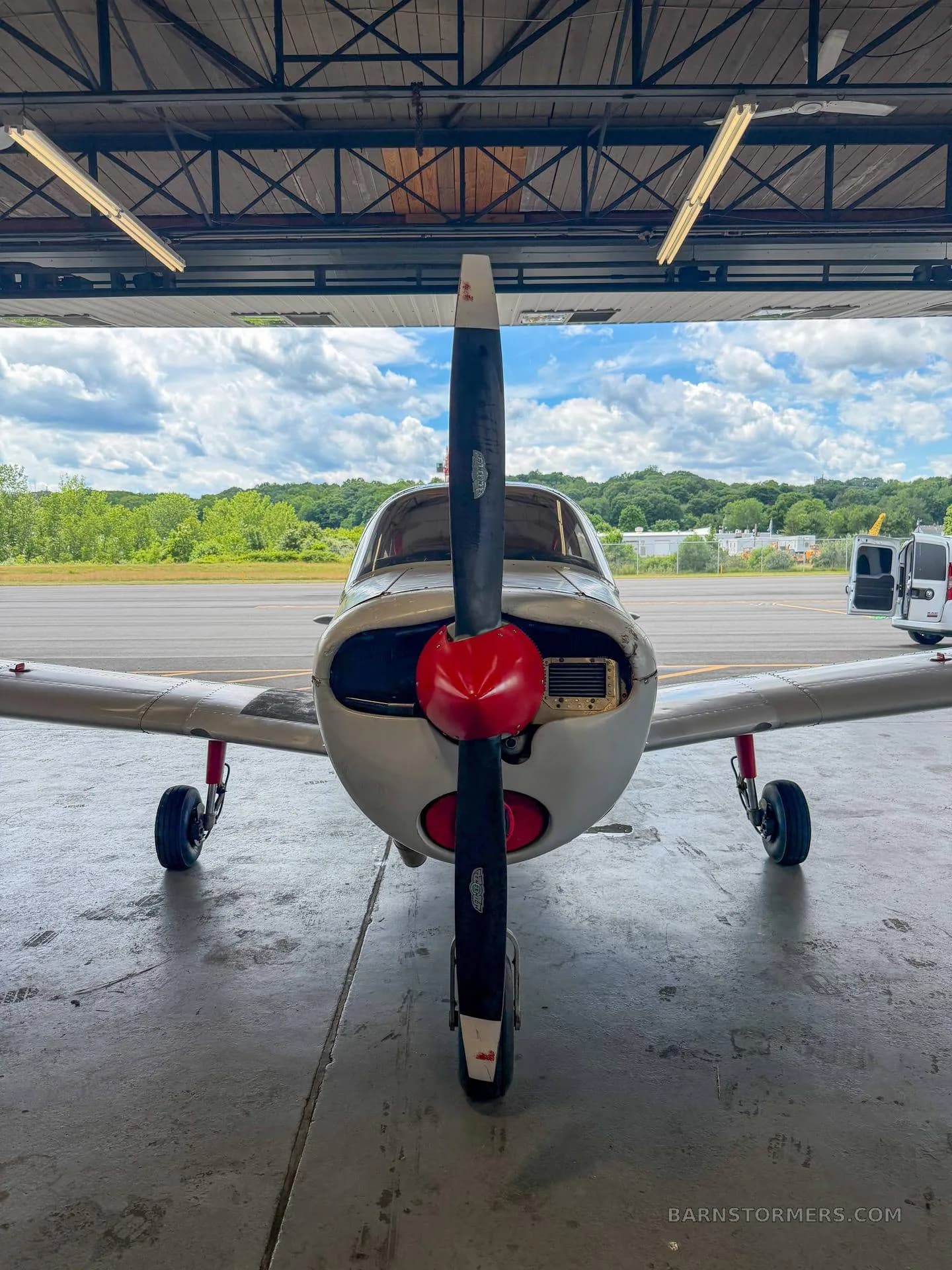 A white single-engine aircraft with a red propeller spinner, viewed from the front, partially inside a hangar with an airfield outside.