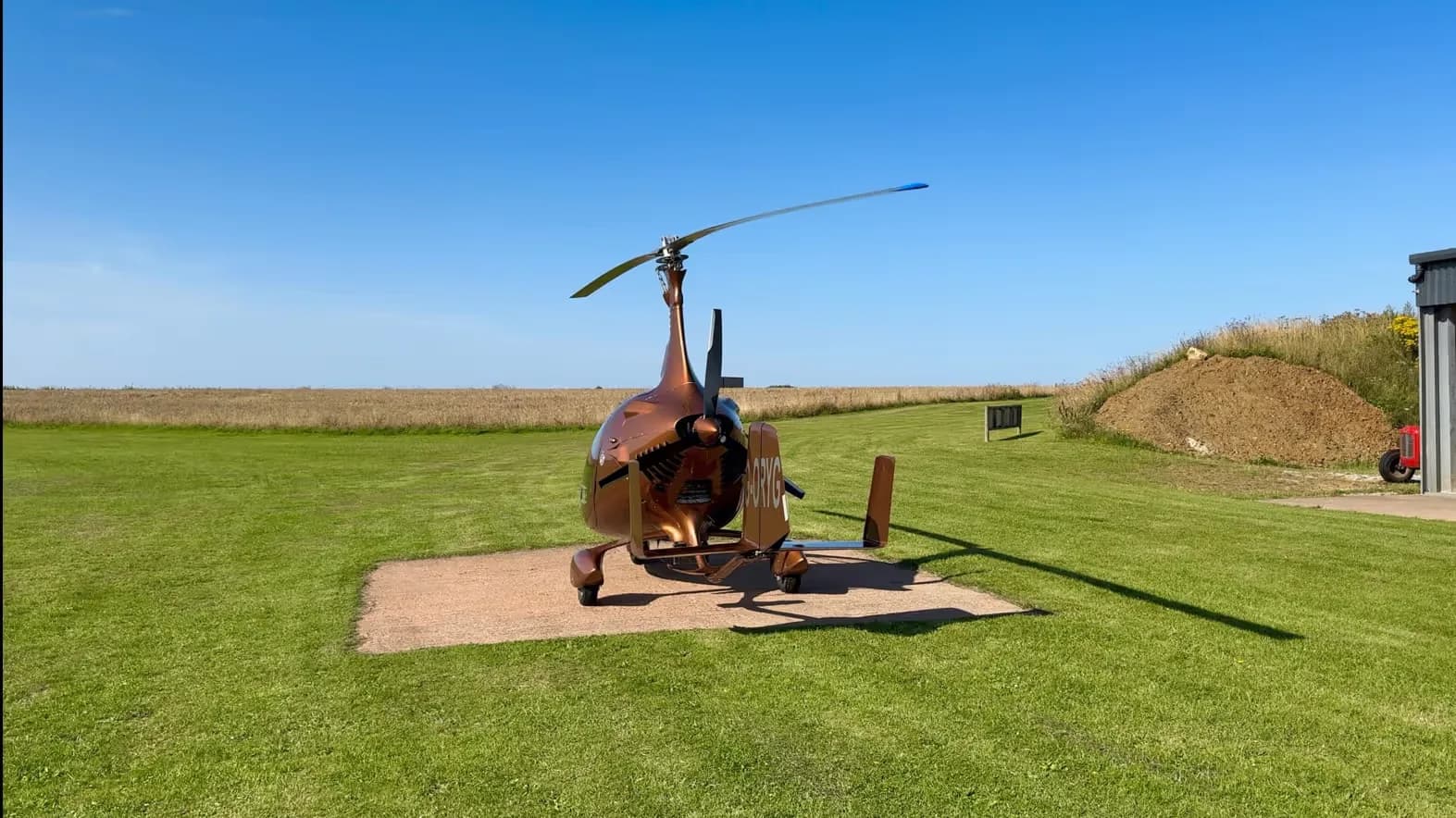 Rear-left view of a brown gyrocopter parked on a concrete pad in a grassy field near a hangar.