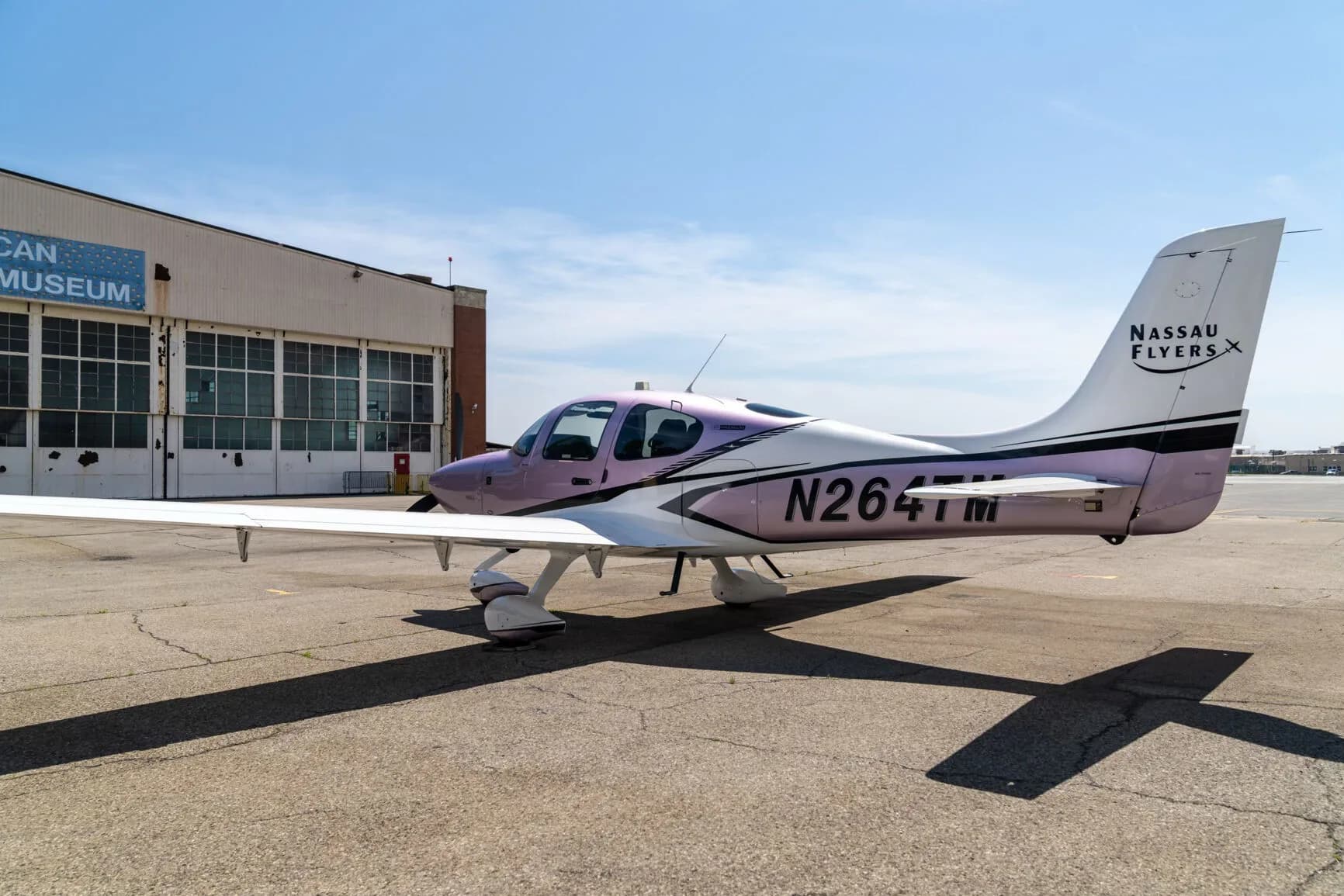 A Cirrus SR22 aircraft with a metallic lavender and white paint scheme, registration N264TM, parked on an airfield next to a building.