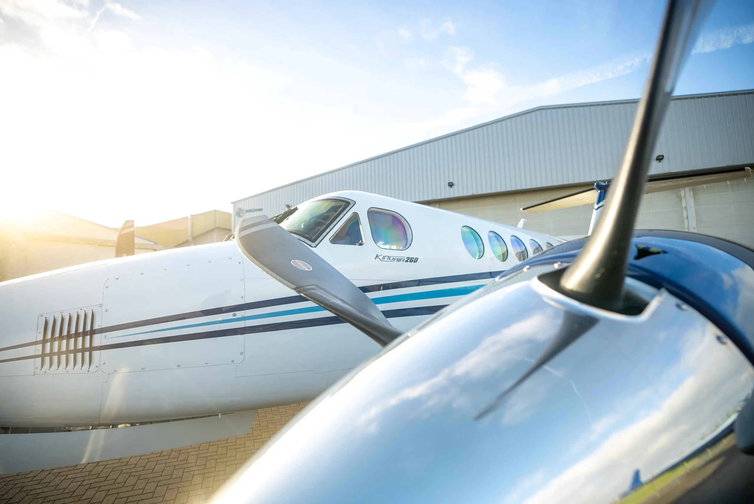 A close-up view of the front fuselage and propeller of a white and blue Beechcraft King Air 260, with sunlight glinting off the propeller.