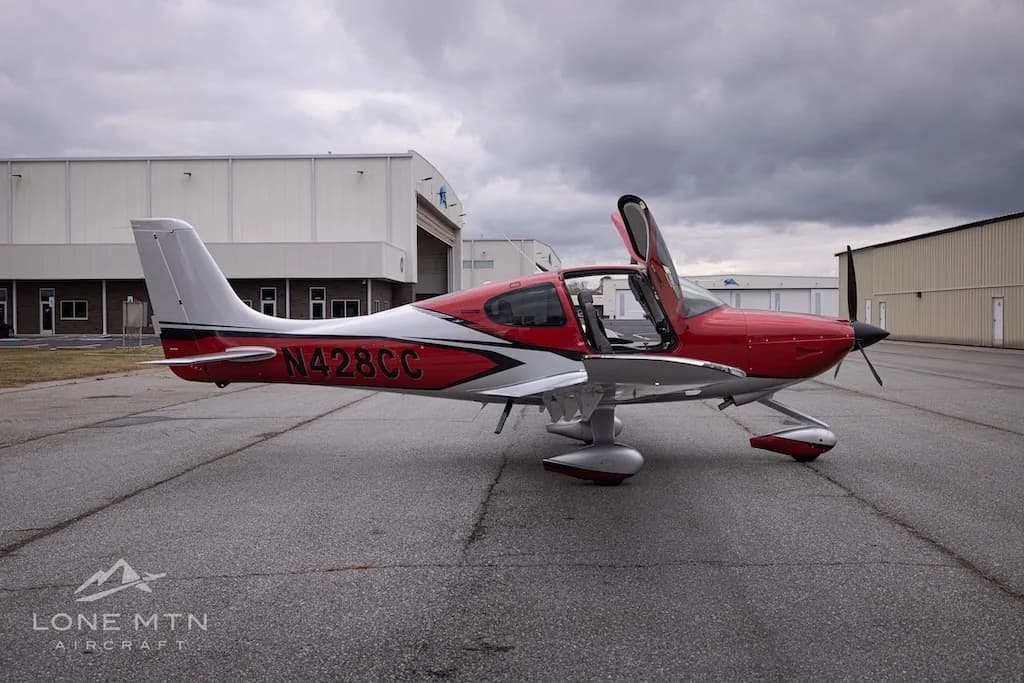 A side view of the red and silver Cirrus Vision Jet (N428CC) with its doors open, parked on an asphalt tarmac.
