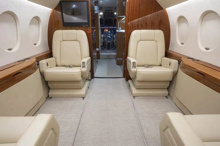 The luxurious interior of a private jet cabin with two cream-colored leather seats facing forward and a view into the cockpit.