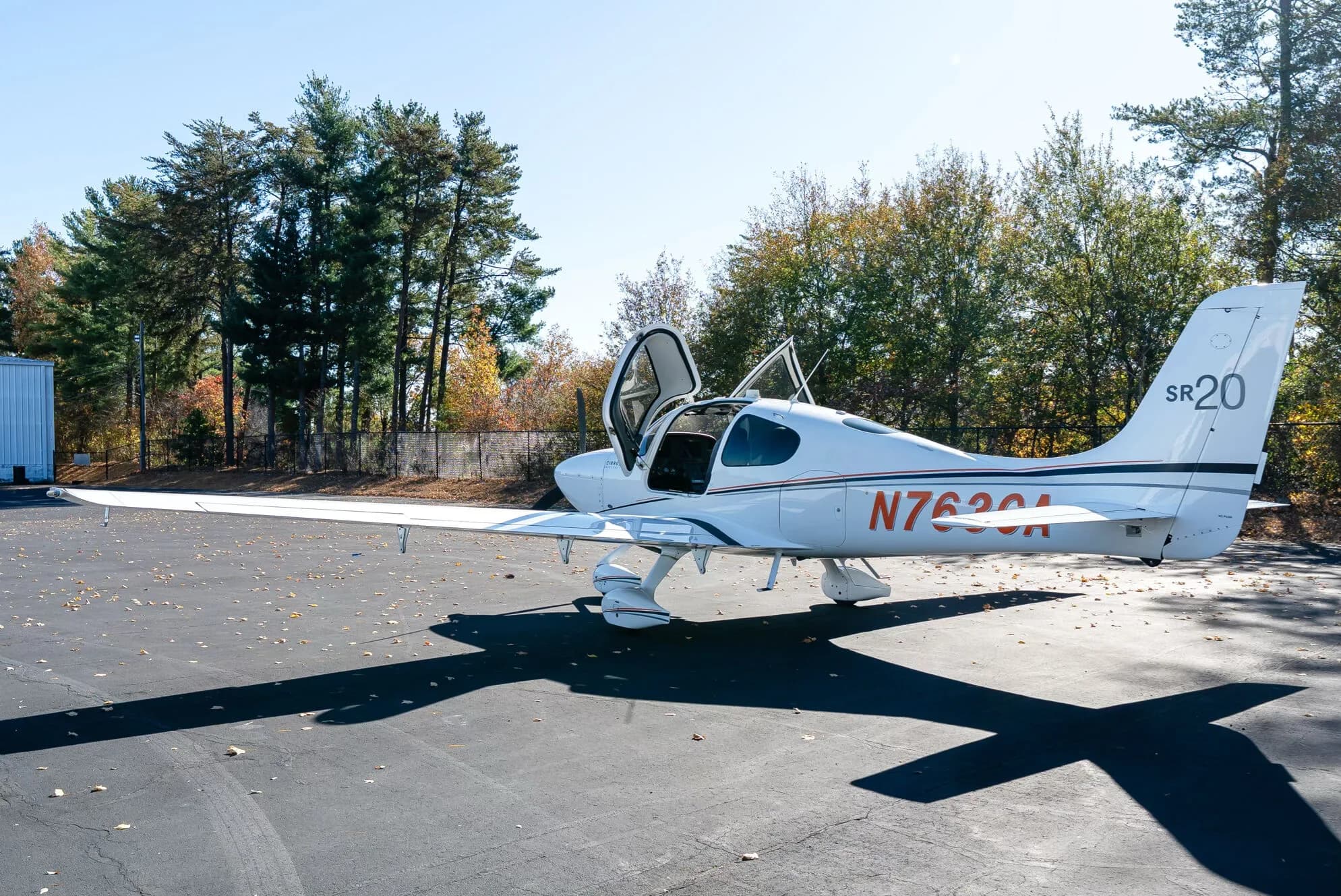 A white Cirrus SR20 aircraft with its doors open is parked on an asphalt tarmac, viewed from the rear left.