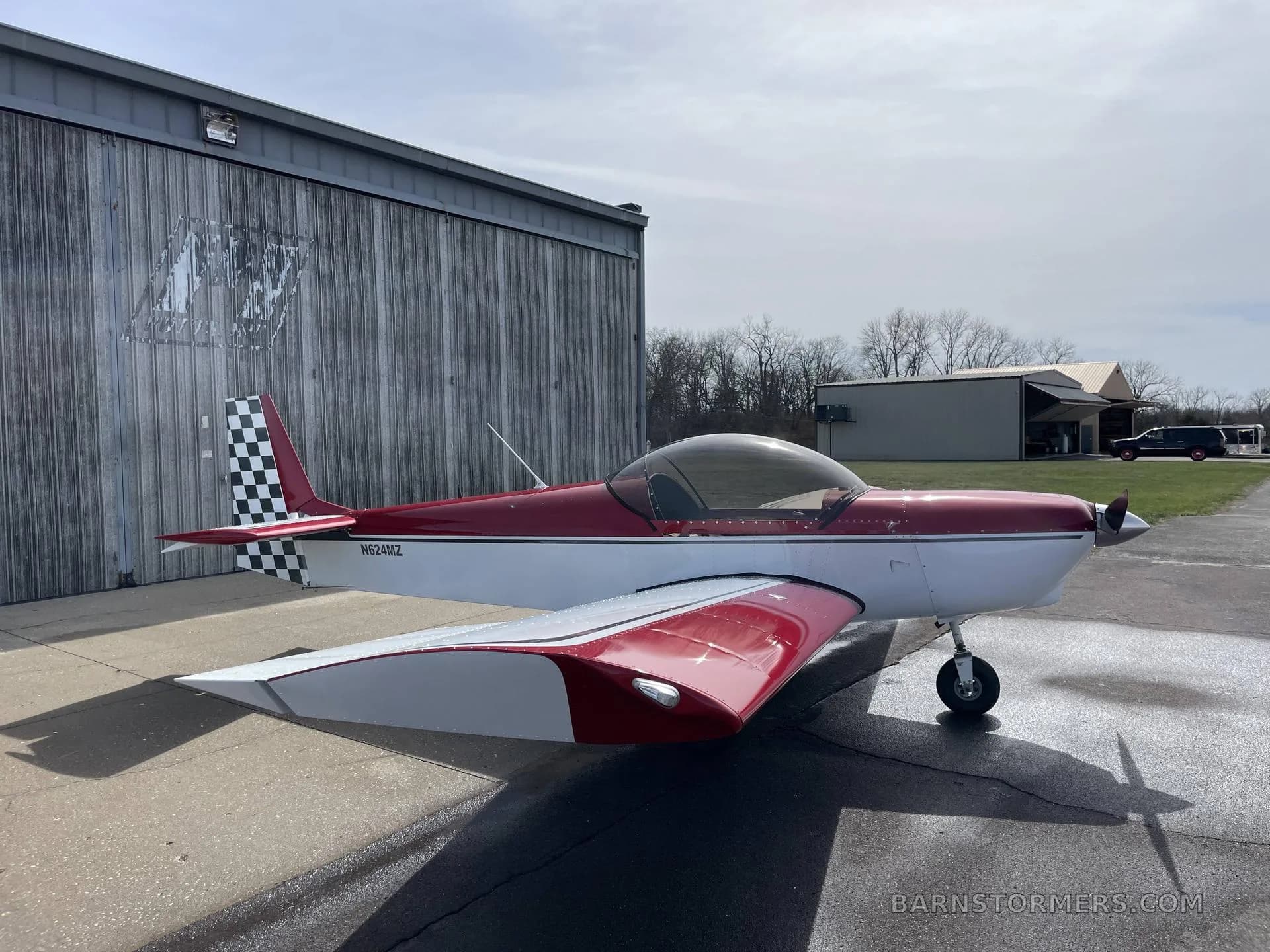 A red and white single-engine aircraft with a checkered tail (N624MZ) is parked on an apron next to a hangar.