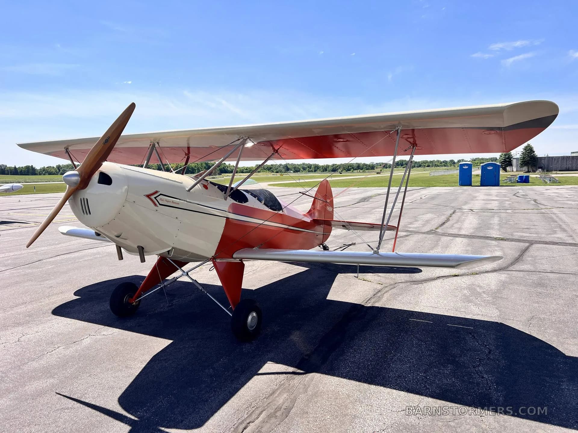 A red and white biplane with a wooden propeller is parked on an asphalt tarmac under a clear blue sky.