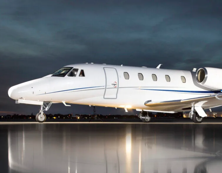 A white business jet with a blue stripe is parked on a wet tarmac at night, reflecting its lights.