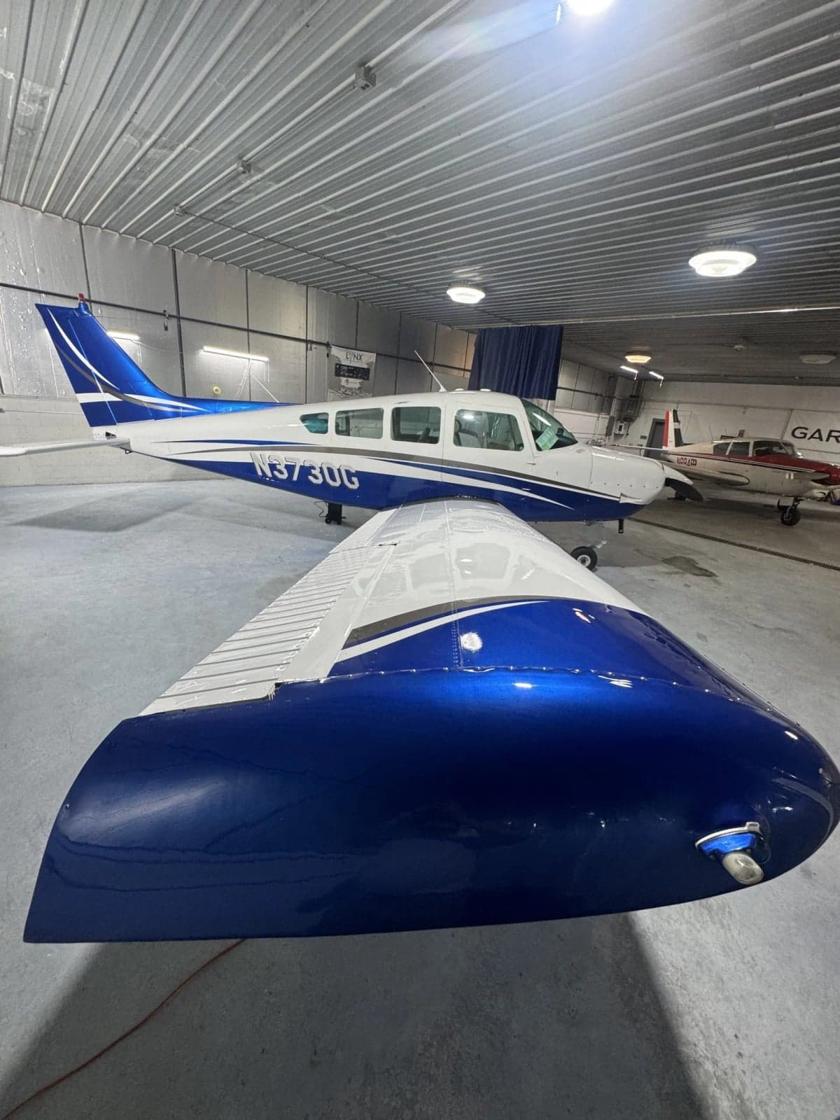 A white and blue single-engine aircraft, N3730G, parked inside a well-lit hangar.