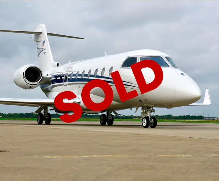 A white business jet with blue stripes is parked on an airfield under a cloudy sky, with 'SOLD' text overlaid.