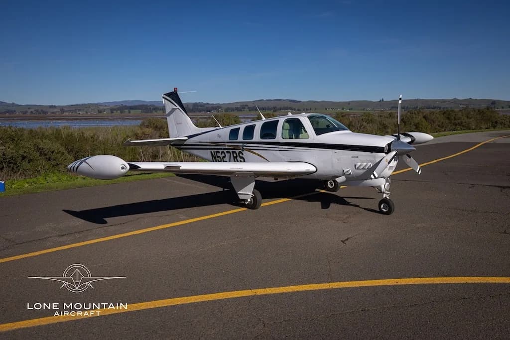A white Beechcraft Baron N527RS with black and gold stripes parked on an asphalt runway with water and hills in the background.