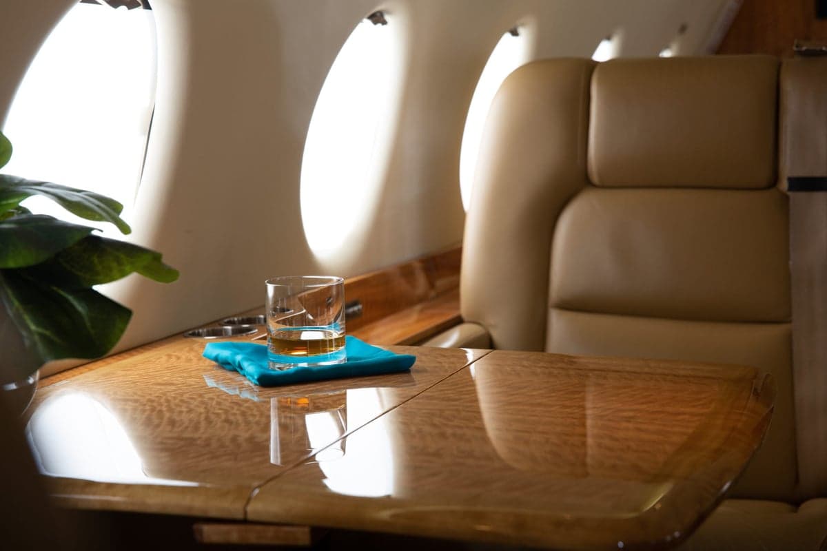 A close-up of a polished wood table inside an aircraft cabin with a glass of whiskey and a beige leather seat.