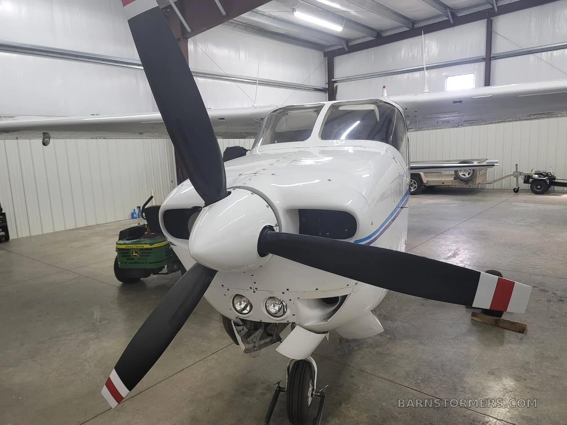 A close-up front view of a white single-engine Cessna aircraft with a three-bladed propeller, parked in a hangar.