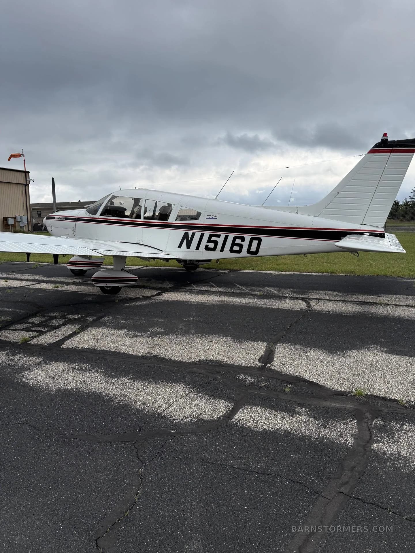 A white Piper Cherokee aircraft with black and red stripes, registration N15160, parked on an asphalt ramp under a cloudy sky.
