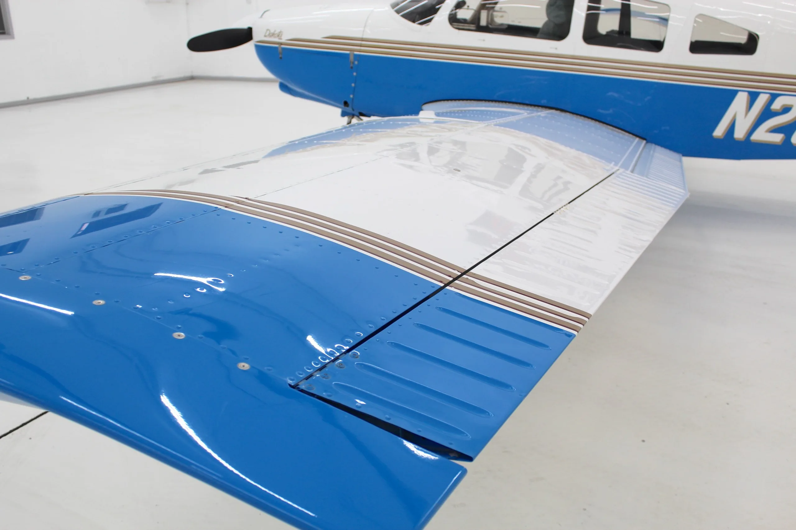 A close-up view of the right wing of the white and blue Piper Dakota aircraft, showing the flaps.