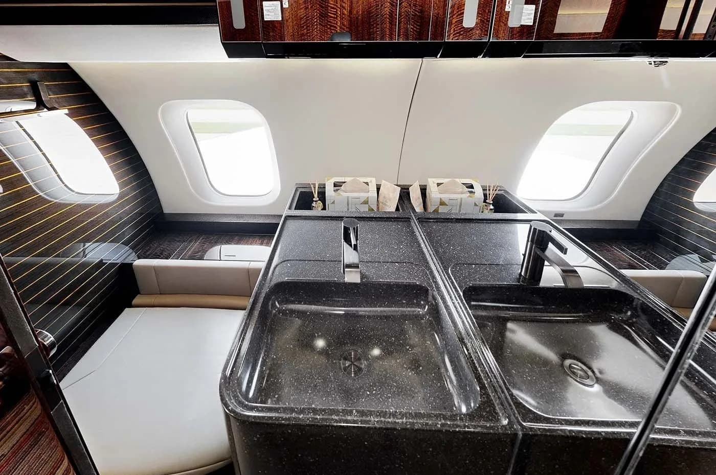 A lavatory or wet bar area in the Bombardier Global business jet, with a black double sink and dark wood accents.