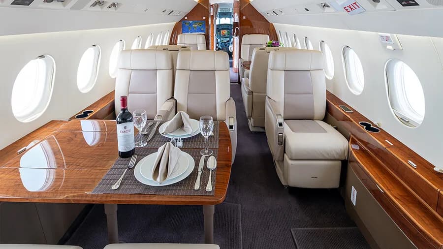 A spacious private jet cabin with a large dining table set for two, surrounded by comfortable beige leather seats and polished wood.