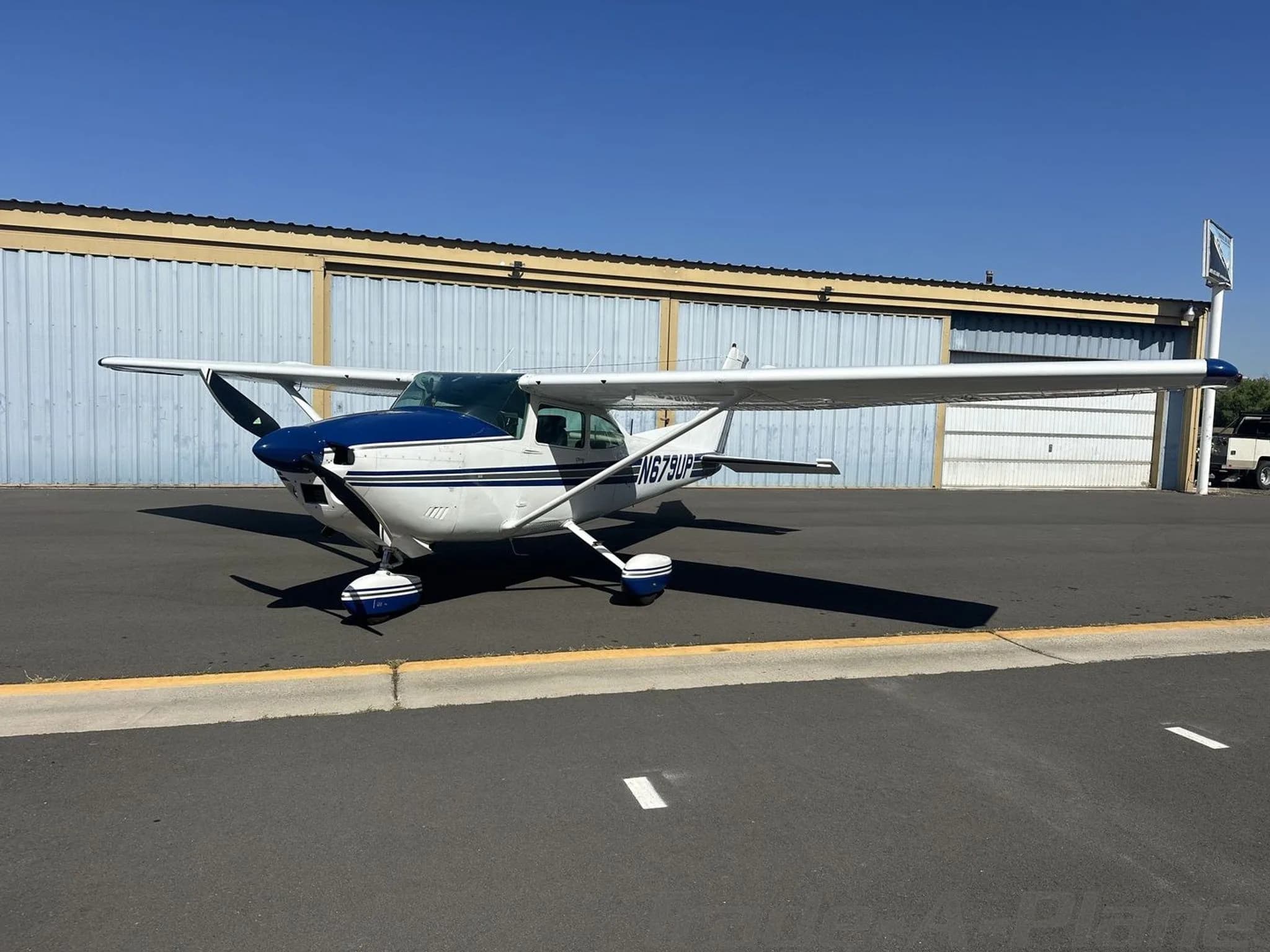 A white and blue Cessna 182 Skylane aircraft parked on an asphalt ramp, viewed from the front-left.