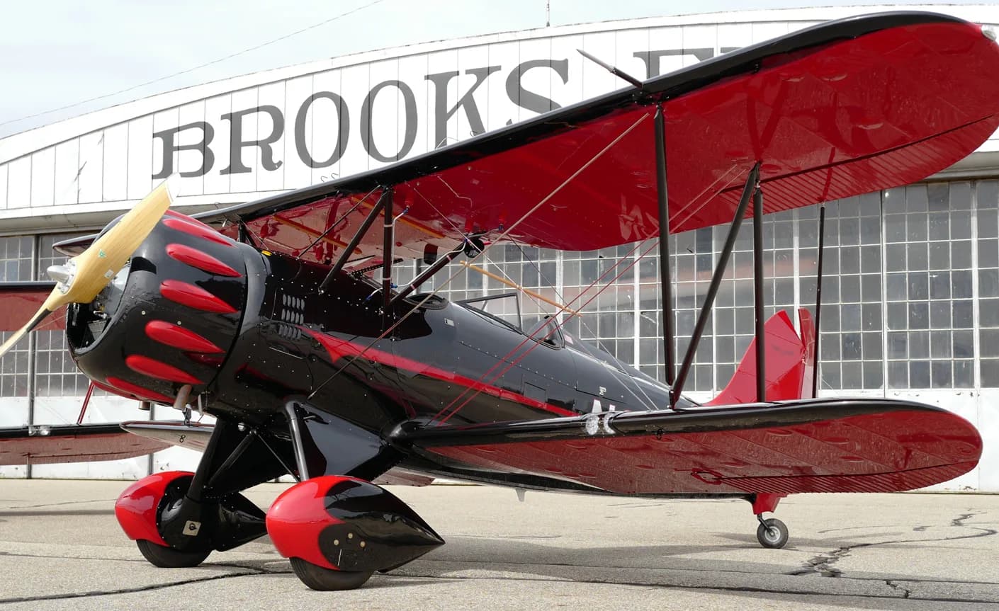 A black and red biplane is parked in front of a hangar with the word 'BROOKS' on it.