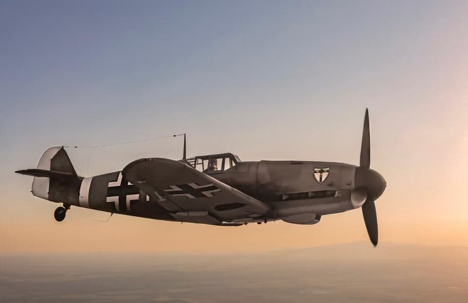 A gray Messerschmitt Bf 109 aircraft in flight over a forest.