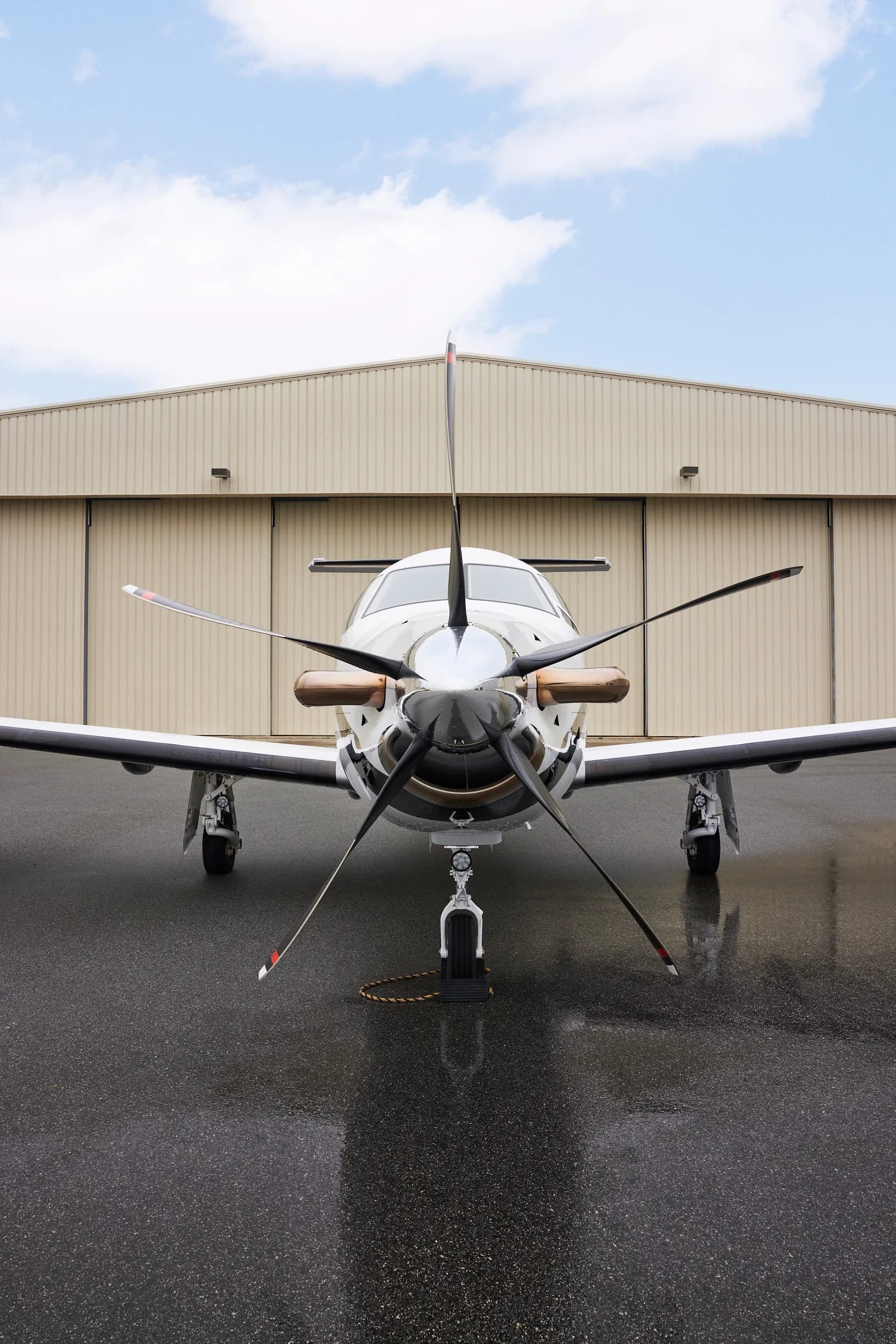 A white Pilatus PC-12 aircraft with black and orange accents parked head-on on a wet tarmac in front of a hangar.