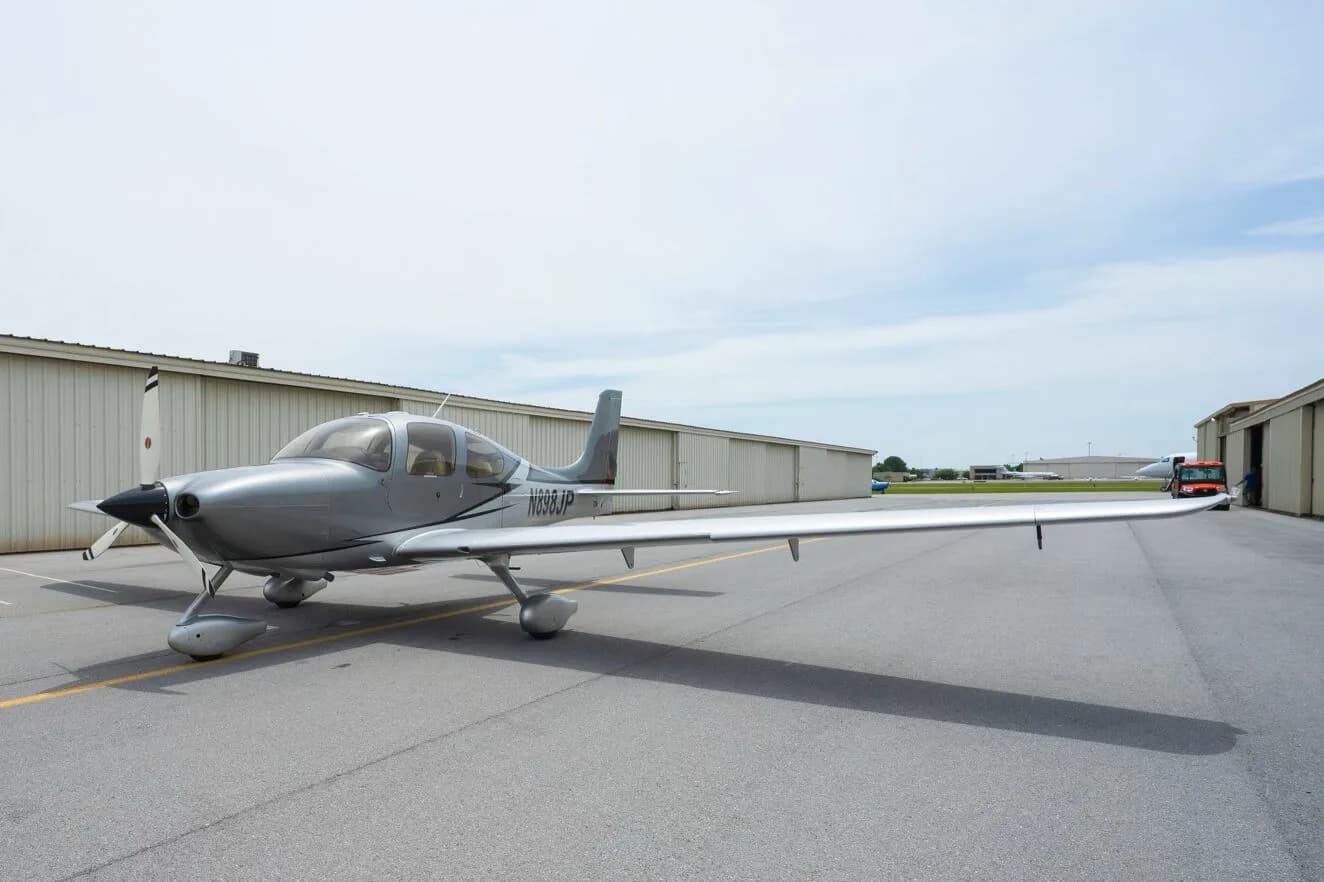 A silver Cirrus SR22 aircraft with registration N898JP, parked on an airfield with hangars in the background.