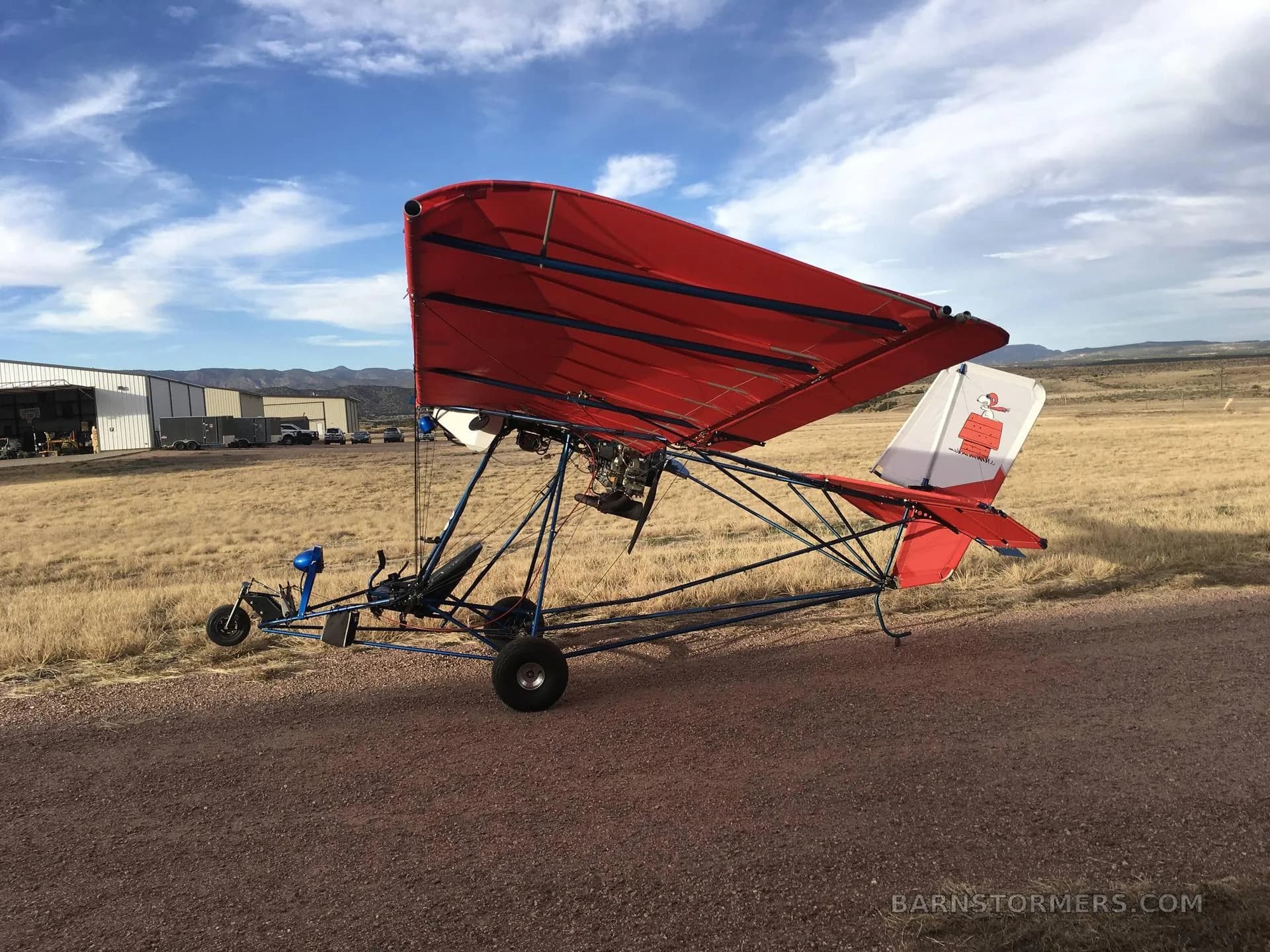 A red ultralight aircraft with a Snoopy tail, viewed from the side on a dirt path under a partly cloudy sky.