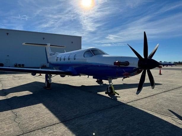 A Pilatus PC-12 aircraft with a blue and white paint scheme is parked on the tarmac in front of a hangar.