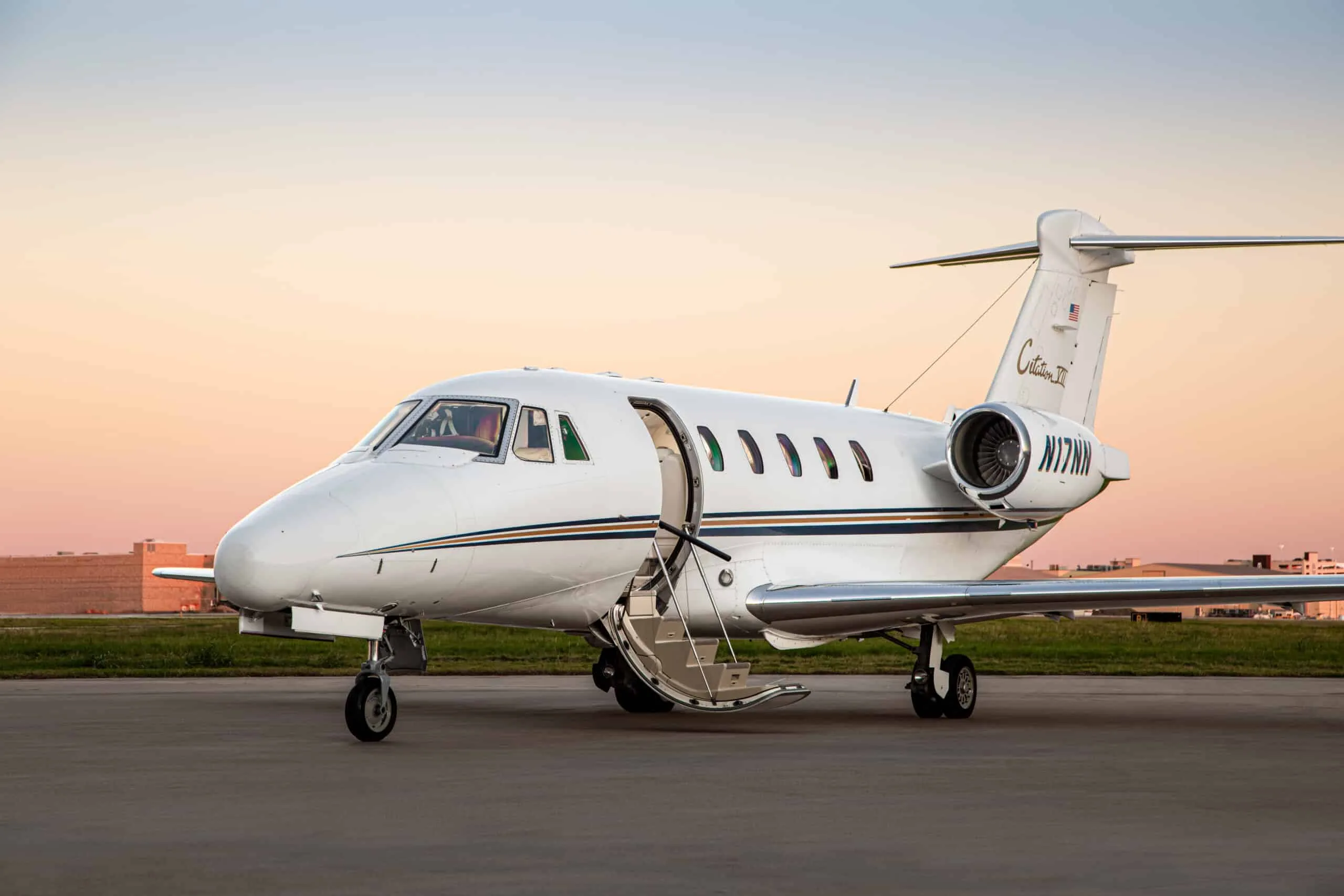 A white Cessna Citation III business jet with tail number N77NN, parked on a tarmac at sunset with its door open.