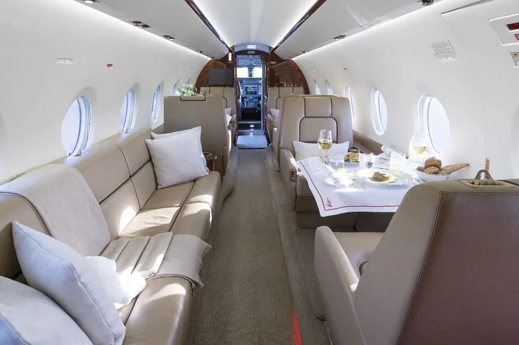 The interior cabin of a private jet with a dining table set for a meal and a long beige leather couch.