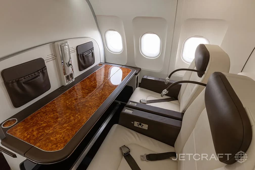 Two individual aircraft seats facing a long wooden table with storage pockets on the wall.