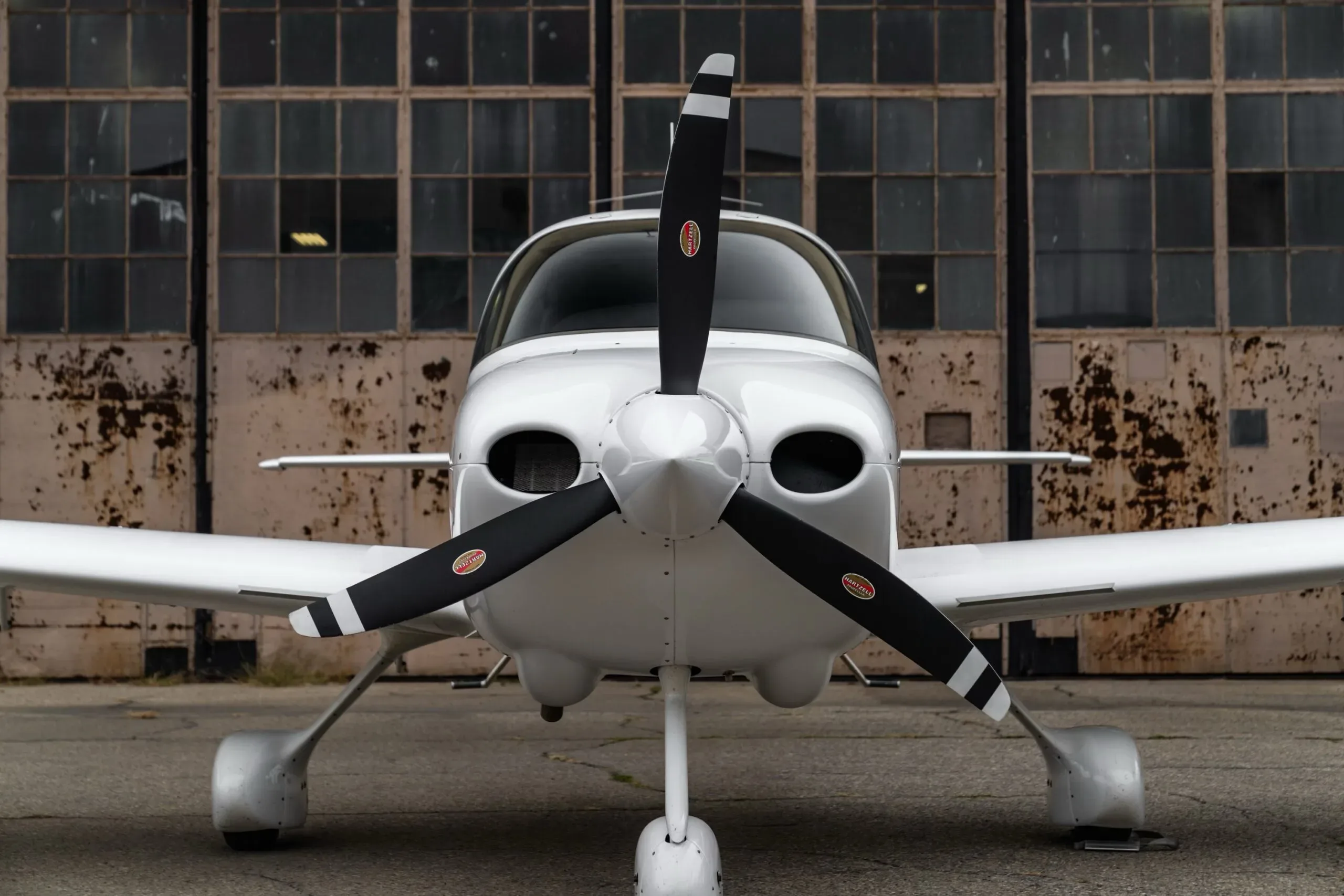 A head-on view of a white Cirrus SR22 aircraft with a three-bladed propeller, parked in front of a weathered hangar.