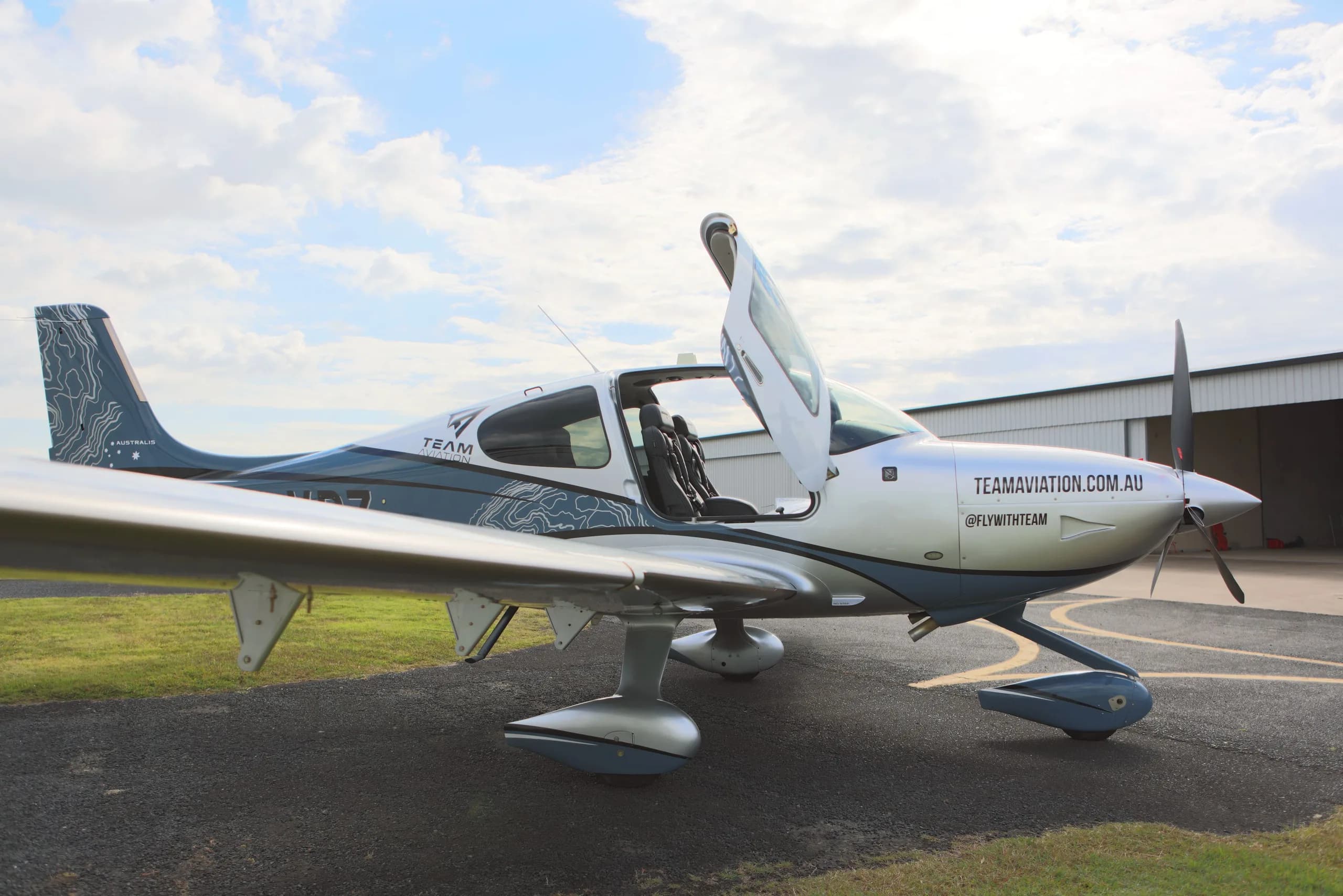A silver and blue single-engine propeller aircraft with open doors and a topographic map paint scheme is parked on an airfield with a hangar in the background.