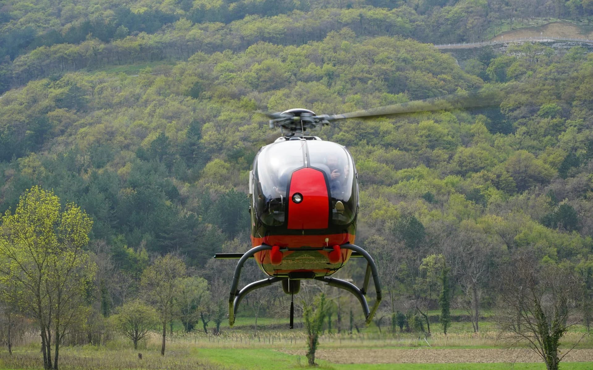 A black and red helicopter is shown from a low angle, with green trees in the background.