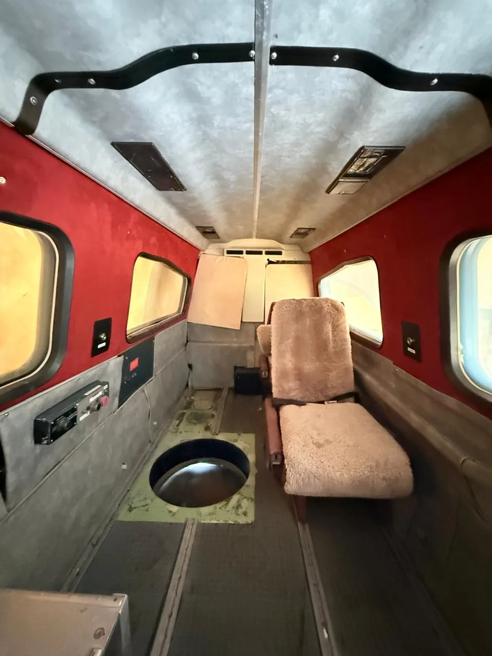 The aircraft's cabin interior has red walls, a single sheepskin-covered seat, and an open circular floor hatch.