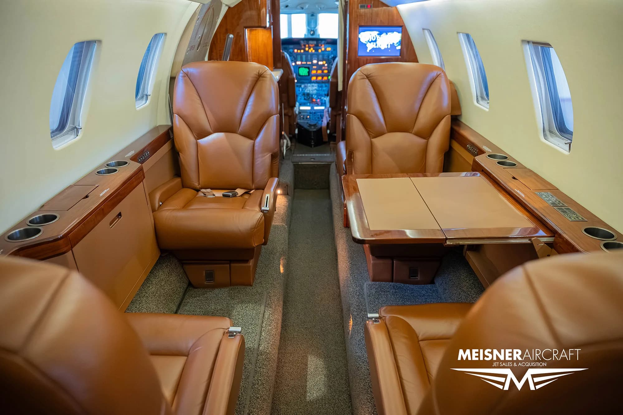 The interior cabin of a private jet featuring brown leather seats and a fold-out table, looking towards the cockpit.