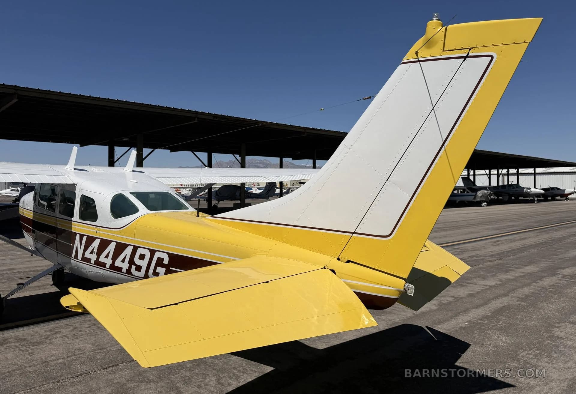 A rear-side view of a white and yellow Cessna 182 aircraft with registration N4449G, showing its tail and right wing, parked on an apron.