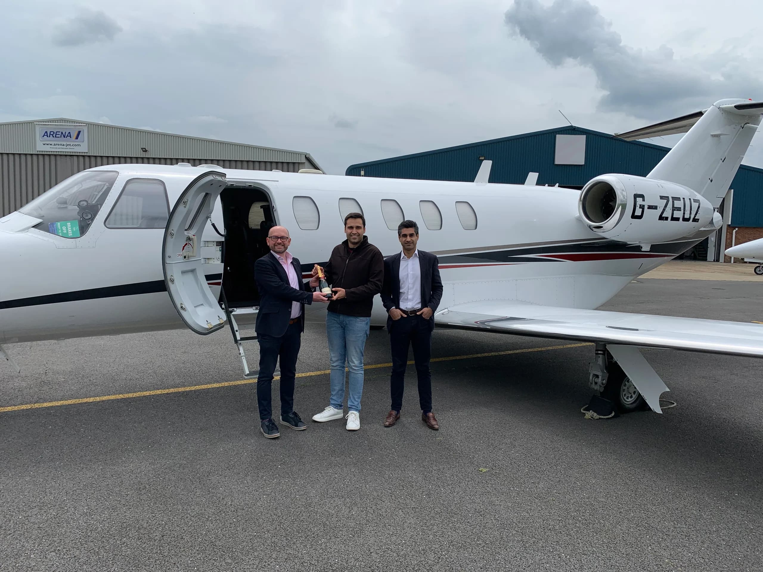 Three men stand next to a white business jet with black, red, and grey stripes, registration G-ZEUZ, on an airport tarmac with a hangar in the background.