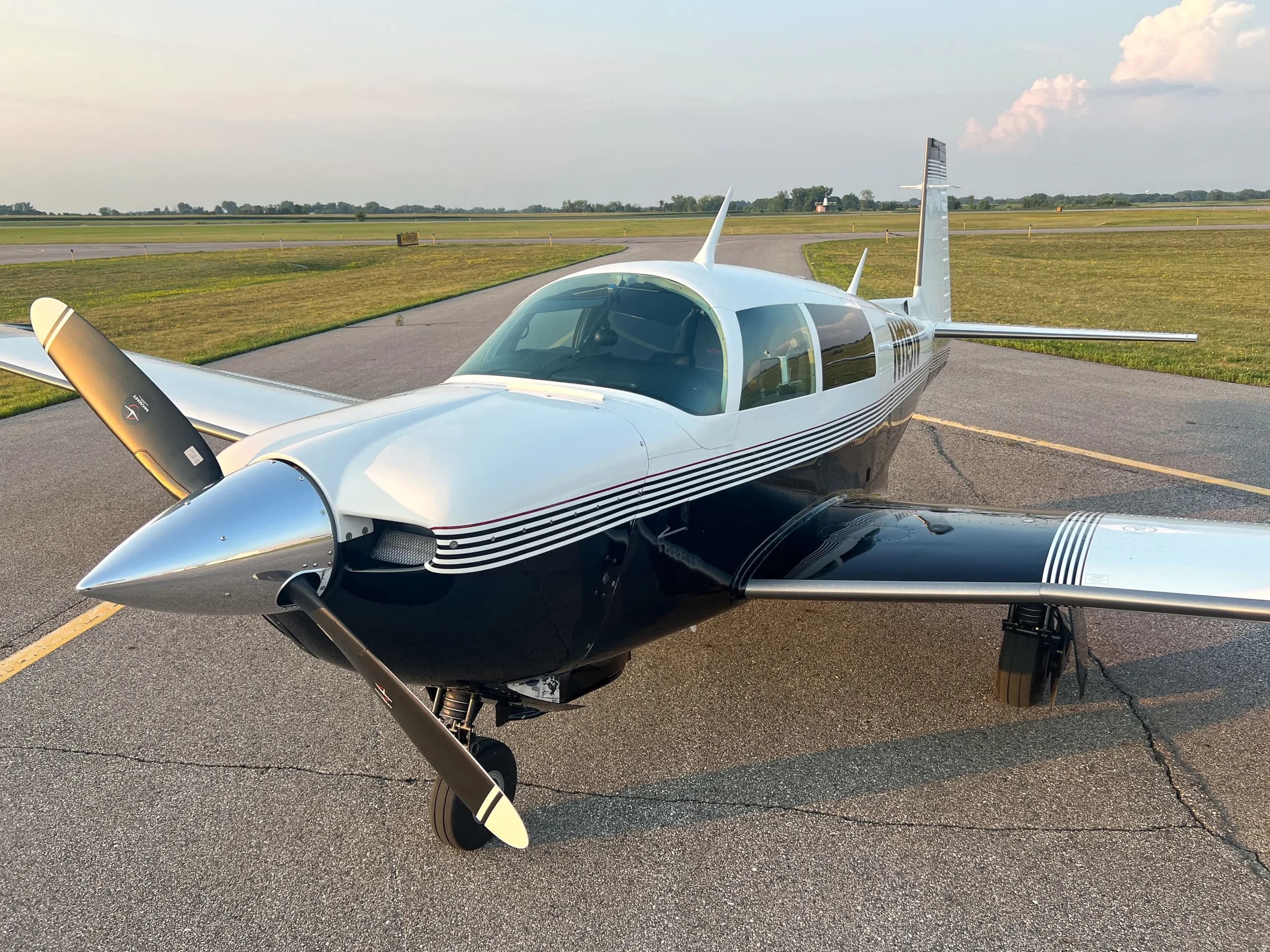 A front-side view of a white and dark blue single-engine aircraft with a polished propeller spinner on an airfield.