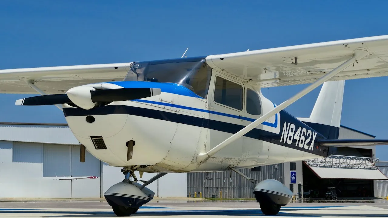A front-right view of a white Cessna 172 with blue and black stripes, N8462X, parked on an airfield near hangars.