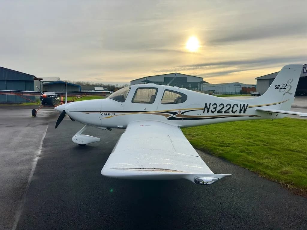 A side view of a white Cirrus SR22 aircraft with N322CW registration, parked on a wet tarmac with the sun low in the sky.