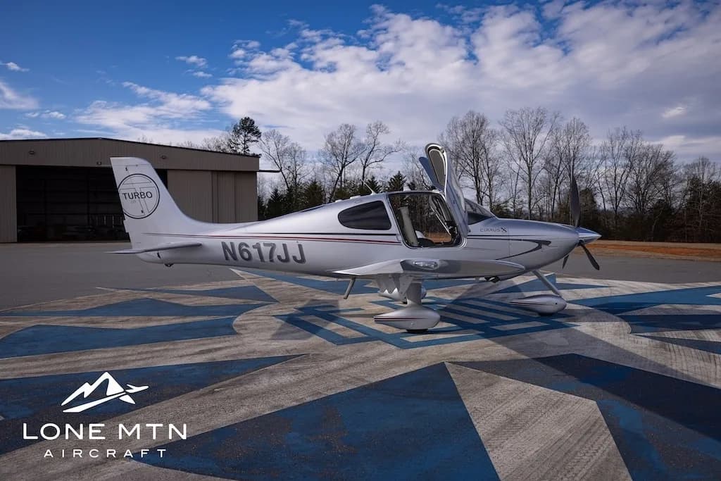 A silver Cirrus SR22T aircraft, N617JJ, with its doors open, parked on an airfield next to a hangar.