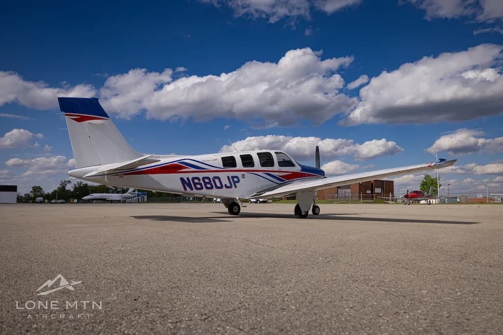 A rear-side view of a white, blue, and red Beechcraft Bonanza N680JP parked on an airfield under a bright blue sky.