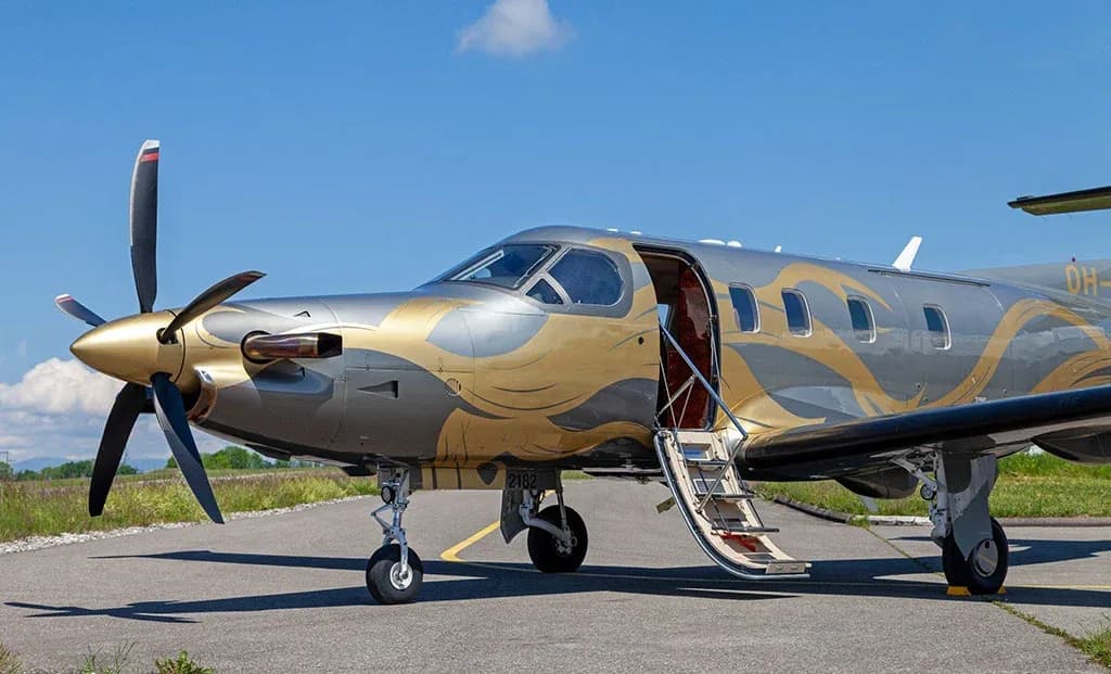 A silver and gold Pilatus PC-12 aircraft with its passenger door open, parked on an asphalt runway under a blue sky.